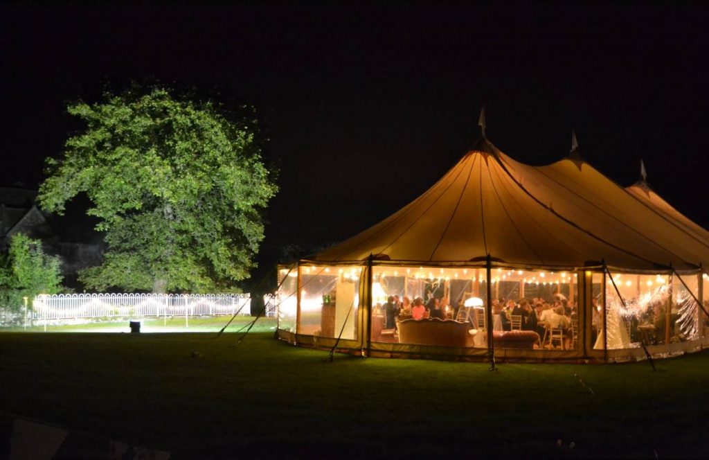 Luxury marquee hire tent illuminated at night for an outdoor wedding or event, featuring warm lighting, seated guests, and a beautifully landscaped garden setting.